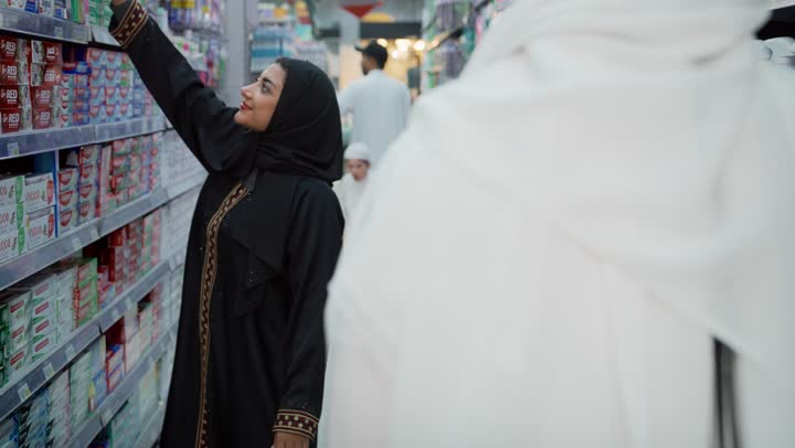 مشاركة الزوج في التبضع وشراء الإحتياجات المنزلية من السوبر ماركت، امراه عربية خليجية إماراتية محجبة ترتدي العباءة تقوم باختيار معجون الأسنان، التأثير الإيجابي للتسوق على العائلة, اختيار بعض الأصناف اللازمة بإيماءات السرور، قضاء وقت ممتع في متجر المواد الغذاائية