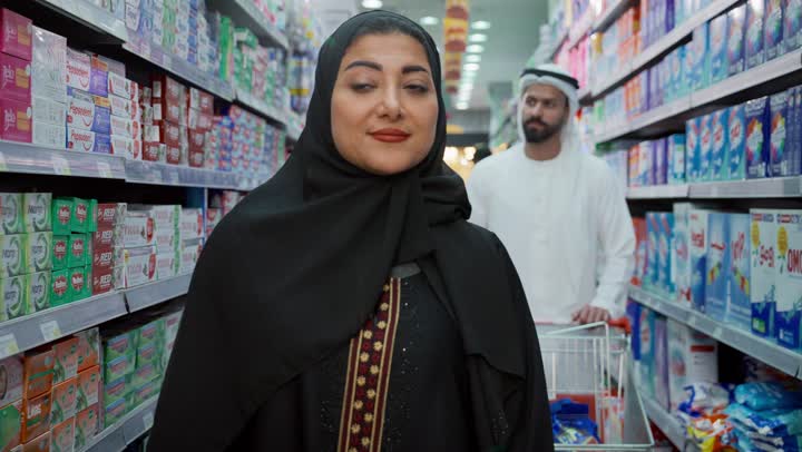 التأثير الإيجابي للتسوق على العائلة، قضاء وقت ممتع في متجر المواد الغذائية، مشاركة الزوج في التبضع وشراء الإحتياجات المنزلية من السوبر ماركت، تصوير مقرب لامرأة عربية خليجية إماراتية محجبة ترتدي العباءة تقوم باختيار اختيار بعض الأصناف اللازمة