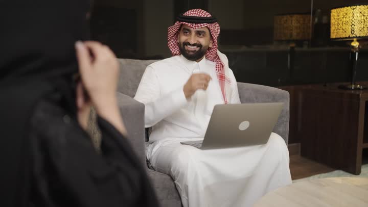 تصفح مواقع التواصل الاجتماعي ، إجراء مكالمة هاتفية عبر الجوال، تصوير امراه عربية خليجية سعودية ترتدي الحجاب والعباءة السوداء تجلس على الأريكة في غرفة المعيشة وتقوم باستخدام الهاتف المحمول ، بجانها رجل يستخدم الحاسوب المحمول .
