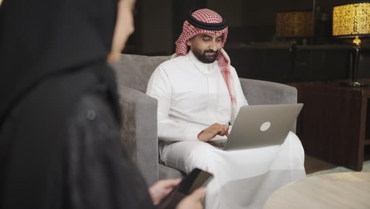 استخدام التكنولوجيا والتقنيات الحديثه ، إجراء مكالمة هاتفية عبر الجوال، تصوير امراه عربية خليجية سعودية ترتدي الحجاب والعباءة السوداء تجلس على الأريكة في غرفة المعيشة وتقوم بالتحدث عبر الهاتف المحمول ، بجانها رجل يستخدم الحاسوب المحمول .