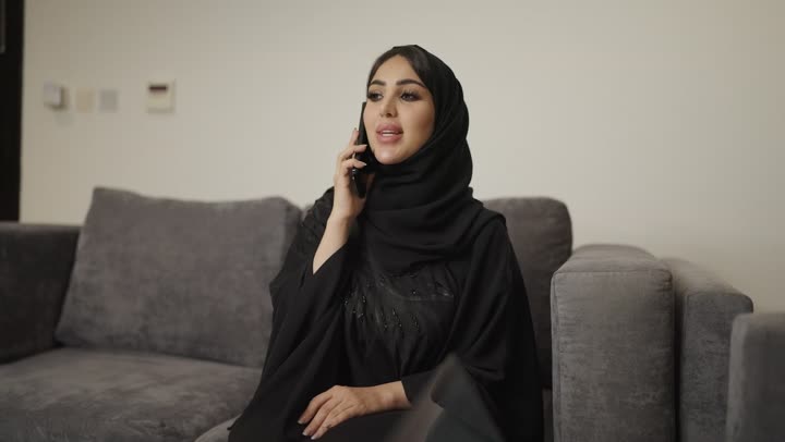 إجراء مكالمة هاتفية عبر الجوال،تصوير امراه عربية خليجية سعودية ترتدي الحجاب والعباءة السوداء تنظر إلى الكاميرا وتجلس على الأريكة في غرفة المعيشة وتقوم بالتحدث عبر الهاتف المحمول ، بجانها رجل يستخدم الحاسوب المحمول .