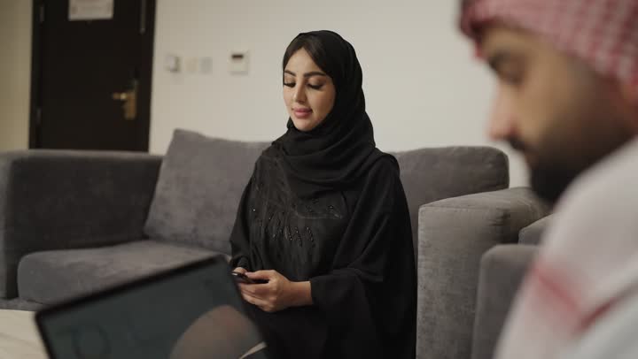 العمل عن بعد وتصفح الانترنت ، إجراء مكالمة هاتفية عبر الجوال، تصوير امراه عربية خليجية سعودية ترتدي الحجاب والعباءة السوداء تجلس على الأريكة في غرفة المعيشة وتقوم باستخدام الهاتف المحمول ، بجانها رجل يستخدم الحاسوب المحمول .