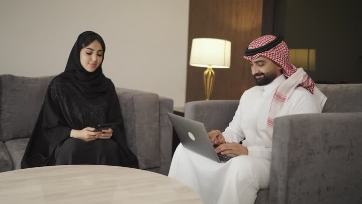 تصفح مواقع التواصل الاجتماعي ، إجراء مكالمة هاتفية عبر الجوال، تصوير امراه عربية خليجية سعودية ترتدي الحجاب والعباءة السوداء تجلس على الأريكة في غرفة المعيشة وتقوم باستخدام الهاتف المحمول ، بجانها رجل يستخدم الحاسوب المحمول .