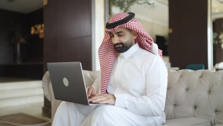 استخدام الأجهزة التقنية الحديثة و المتطورة ,أداء المهام والاعمال المكتبية،رجل عربي خليجي سعودي يجلس على الاريكة في غرفة فندقية، استخدام جهاز الحاسوب المحمول، العمل عن بعد وتصفح الانترنت، انجاز مهام العمل، احتراف وظائف الحاسوب وتكنولوجيا المعلومات