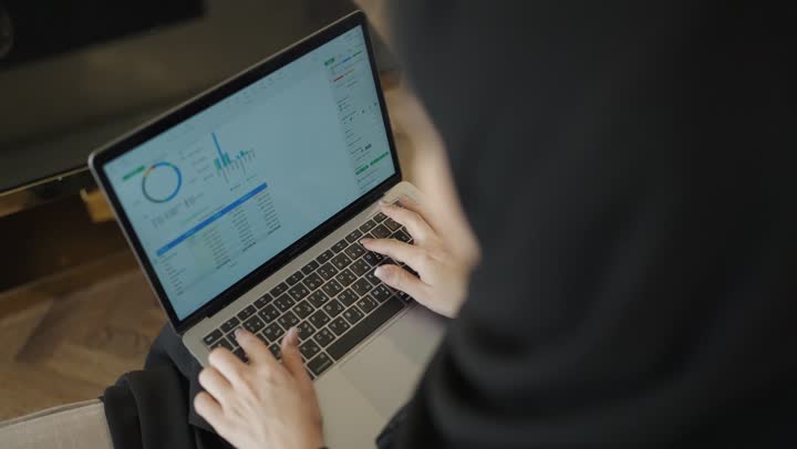 تصوير لامرأة عربية خليجية سعودية ترتدي العباءة والحجاب تجلس على الاريكة في غرفة فندقية ، استخدام الأجهزة التقنية الحديثة و المتطورة ، استخدام جهاز الحاسوب المحمول، أداء المهام والاعمال المكتبية، احتراف وظائف الحاسوب وتكنولوجيا المعلومات ، العمل عن بعد وتصفح الانترنت