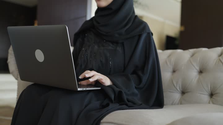 أداء المهام والاعمال المكتبية، احتراف وظائف الحاسوب وتكنولوجيا المعلومات ، العمل عن بعد وتصفح الانترنت، استخدام الأجهزة التقنية الحديثة و المتطورة ،تصوير لامرأة عربية خليجية سعودية ترتدي العباءة والحجاب تجلس على الاريكة في غرفة فندقية، استخدام جهاز الحاسوب المحمول، انجاز مهام العمل،