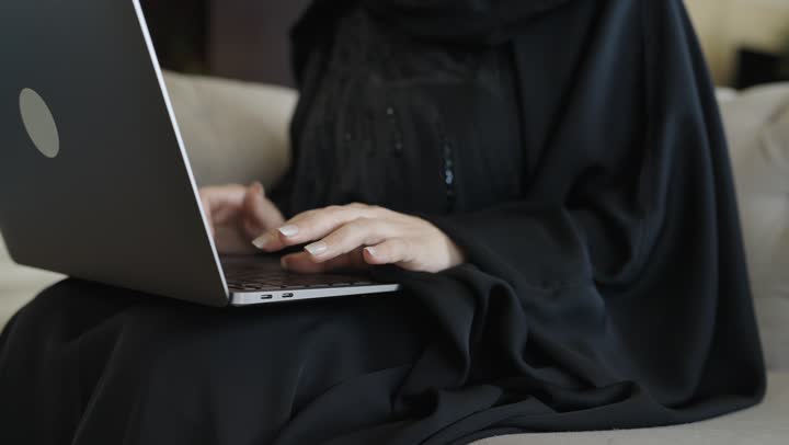 العمل عن بعد وتصفح الانترنت، أداء المهام والاعمال المكتبية،استخدام الأجهزة التقنية الحديثة و المتطورة , احتراف وظائف الحاسوب وتكنولوجيا المعلومات ،تصوير لامرأة عربية خليجية سعودية ترتدي العباءة والحجاب تجلس على الاريكة في غرفة فندقية، استخدام جهاز الحاسوب المحمول، انجاز مهام العمل،