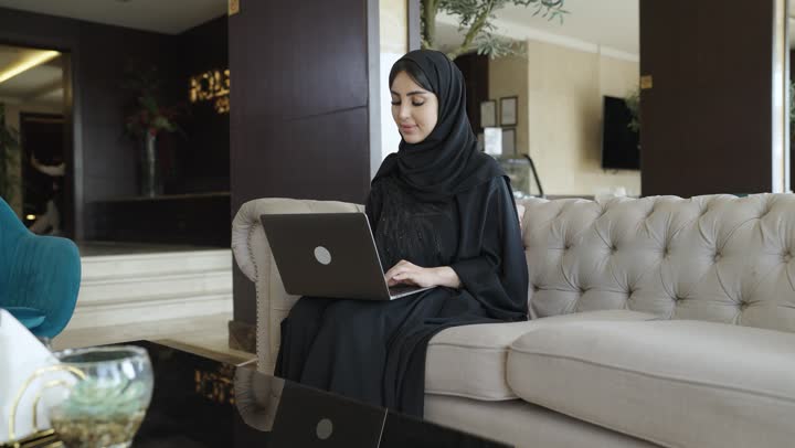 احتراف وظائف الحاسوب وتكنولوجيا المعلومات ، العمل عن بعد وتصفح الانترنت، أداء المهام والاعمال المكتبية،استخدام الأجهزة التقنية الحديثة و المتطورة ,تصوير لامرأة عربية خليجية سعودية ترتدي العباءة والحجاب تجلس على الاريكة في غرفة فندقية، استخدام جهاز الحاسوب المحمول، انجاز مهام العمل،