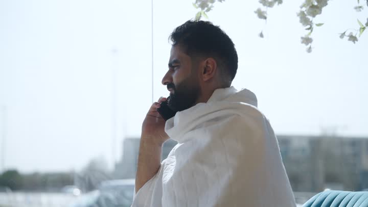 Using modern technical devices to browse guidance sites, using the electronic Hajj and Umrah guide application on the mobile phone, performing Hajj and Umrah rituals, spending enjoyable times outside, a Saudi Arab pilgrim wearing Ihram clothes sitting in an upscale coffee shop holding a mobile phone in his hand