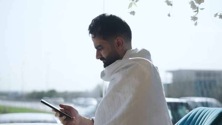 استخدام تطبيق دليل الحاج والمعتمر الالكتروني على الهاتف المحمول, أداء مناسك الحج والعمرة  ، استخدام الأجهزة التقنية الحديثة في تصفح مواقع الارشادات ، قضاء اوقات ممتعه فى الخارج ، حاج عربي سعودي يرتدي ملابس الاحرام يجلس فى كوفى شوب راقي يحمل بيده جهاز الهاتف المحمول