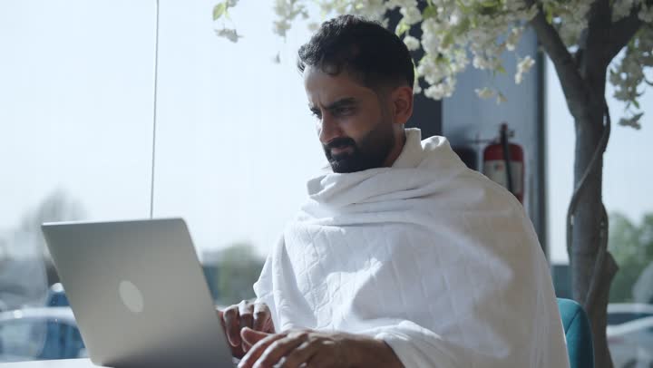 Online booking for Hajj and Umrah campaigns, performing Hajj and Umrah rituals, using modern technical devices to browse guidance websites, spending enjoyable times abroad, a Saudi Arabian pilgrim wearing Ihram clothes sitting in an upscale coffee shop holding a laptop in his hand