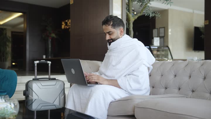 استخدام الأجهزة التقنية الحديثة في تصفح مواقع الارشادات لأداء مناسك الحج والعمرة، حاج عربي سعودي يرتدي ملابس الاحرام يجلس فى مكتب الاستقبال بالفندق يحمل بيده جهاز الحاسوب المحمول بإيماءات السرور