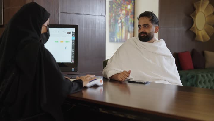 خدمة ومكتب إستقبال العملاء  ، حجز غرفة فندقيه عند مكتب الاستقبال ، طلب الاستشاره والمساعده ، حاج عربي سعودي يرتدي ملابس الاحرام يجلس في مكتب خدمة الحجاج يتحدث الى امرأه عربيه ترتدي عباءه خليجيه وكمامه سوداء ، الاحترازات الوقايه ضد فايروس كورونا