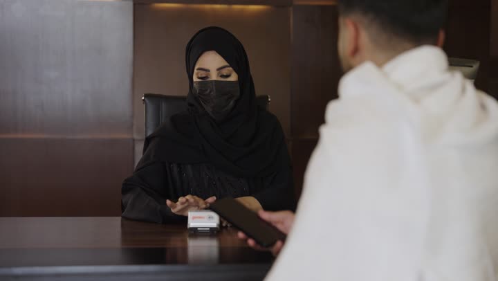 الدفع عبر بطاقه الائتمان، حجز غرفة فندقيه عند مكتب الاستقبال ، طلب الاستشاره والمساعده ، حاج عربي سعودي يرتدي ملابس الاحرام يجلس في مكتب خدمة الحجاج يتحدث الى امرأه عربيه ترتدي عباءه خليجيه وكمامه سوداء ، الاحترازات الوقايه ضد فايروس كورونا