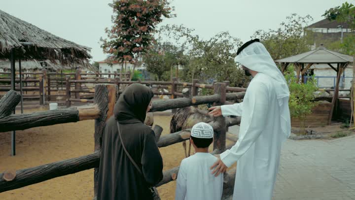 نزهه عائلية في حديقة الحيوانات، تصوير من الخلف لزوجان عربيان خليجيان اماراتيان يرتديان الزي التقليدي يقفان امام قفص طيور النعام برفقه طفلهما، مشاهده الحيوانات و التعرف عليها، اجواء تعليميه و ترفيهيه في الهواء الطلق