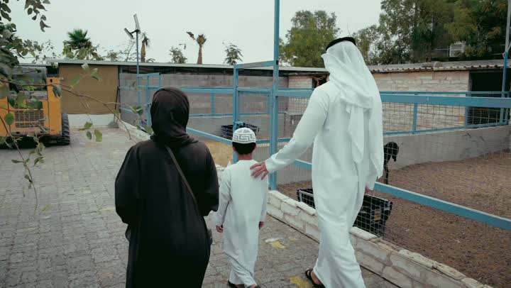 مفهوم الترفيه العائلى، قضاء وقت ممتع و مشاهده الحيوانات، زياره عائلية لحديقة الحيوانات، جوله ترفيهيه وممتعه وسط الاقفاص المفتوحه، تصوير من الخلف لزوجان عربيان خليجان اماراتيان يرتديان الزي التقليدي يقومان بنزهه عائلية خارجيه برفقه طفلهما