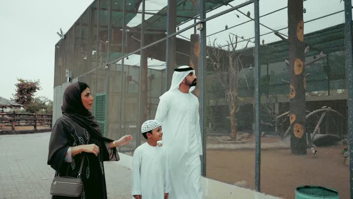 زياره عائلية لحديقة الحيوانات، جوله ترفيهيه وممتعه وسط الاقفاص المفتوحه، زوجان عربيان خليجان اماراتيان يرتديان الزي التقليدي يقومان بنزهه عائلية خارجيه برفقه طفلهما، قضاء وقت ممتع و مشاهده الحيوانات