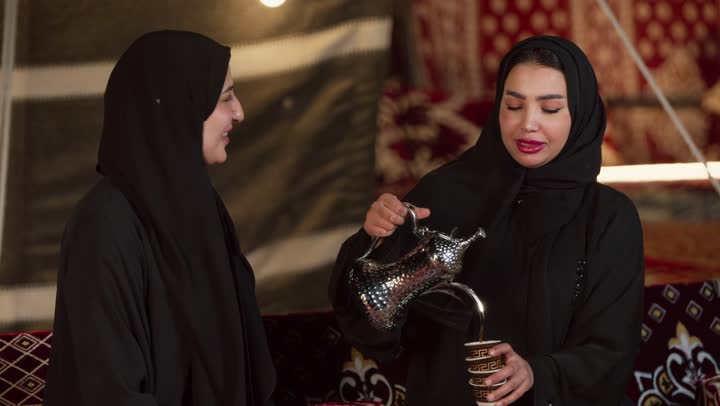 كرم الضيافة وحسن الاستقبال في شهر رمضان, زينة وإضاءة رمضانية، تقديم القهوة العربية الساخنة للضيف، كشتة بر وتخييم في صحراء الإمارات، حوار ونقاش بين الاصدقاء، سيدتان عربيتان اماراتيتان خليجيتان ترتديان العباءة تجلسان في خيمة شعبية