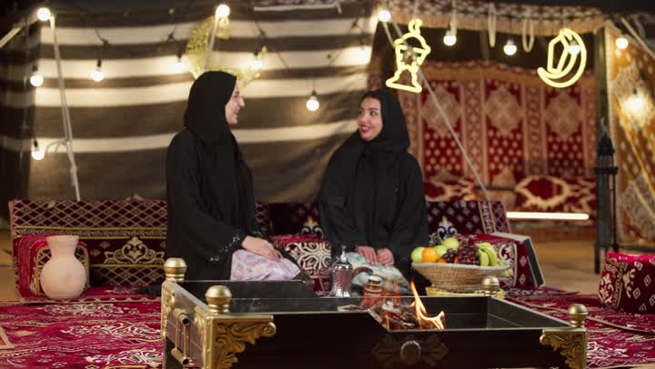 حسن الاستقبال وكرم الضيافة في شهر رمضان, زينة وإضاءة رمضانية، امرأة تقوم بالإشارة بإصبع السبابة إلى شيء ما، كشتة بر وتخييم في صحراء الإمارات، حوار ونقاش بين الاصدقاء بإيماءات السعادة، سيدتان عربيتان اماراتيتان خليجيتان ترتديان العباءة تجلسان في خيمة شعبية