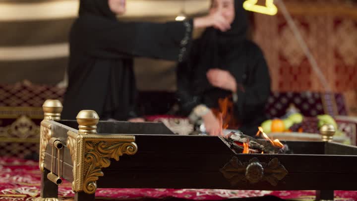 حوار ونقاش بين الاصدقاء بإيماءات السعادة والسرور، الإشارة بإصبع السبابة إلى شيء ما، زينة وإضاءة رمضانية، كشتة بر وتخييم في صحراء الإمارات، سيدتان عربيتان اماراتيتان خليجيتان ترتديان العباءة تجلسان في خيمة شعبية تقليدية