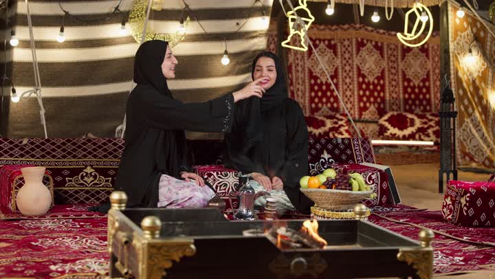 حسن الاستقبال وكرم الضيافة في شهر رمضان, زينة وإضاءة رمضانية، امرأة تقوم بالإشارة بإصبع السبابة إلى شيء ما، كشتة بر وتخييم في صحراء الإمارات، حوار ونقاش بين الاصدقاء بإيماءات السعادة، سيدتان عربيتان اماراتيتان خليجيتان ترتديان العباءة تجلسان في خيمة شعبية