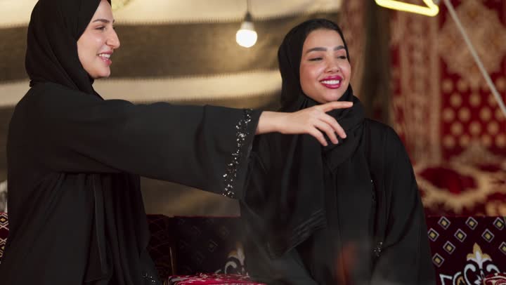 حوار ونقاش بين الاصدقاء بإيماءات السعادة والسرور، الإشارة بإصبع السبابة إلى شيء ما، زينة وإضاءة رمضانية، كشتة بر وتخييم في صحراء الإمارات، سيدتان عربيتان اماراتيتان خليجيتان ترتديان العباءة تجلسان في خيمة شعبية تقليدية