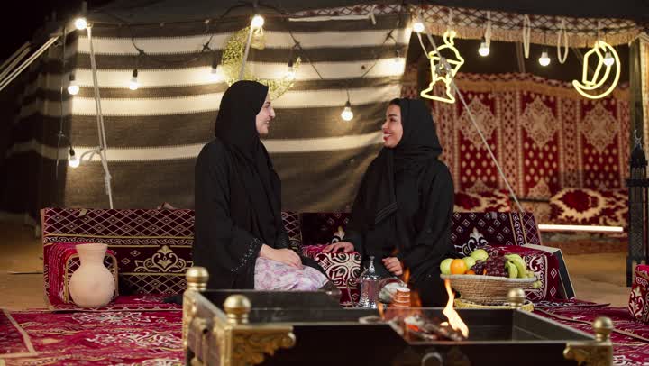 الإشارة بإصبع السبابة إلى شيء ما، حسن الاستقبال وكرم الضيافة في شهر رمضان, زينة وإضاءة رمضانية، كشتة بر وتخييم في صحراء الإمارات، حوار ونقاش بين الاصدقاء بإيماءات السعادة، سيدتان عربيتان اماراتيتان خليجيتان ترتديان العباءة تجلسان في خيمة شعبية