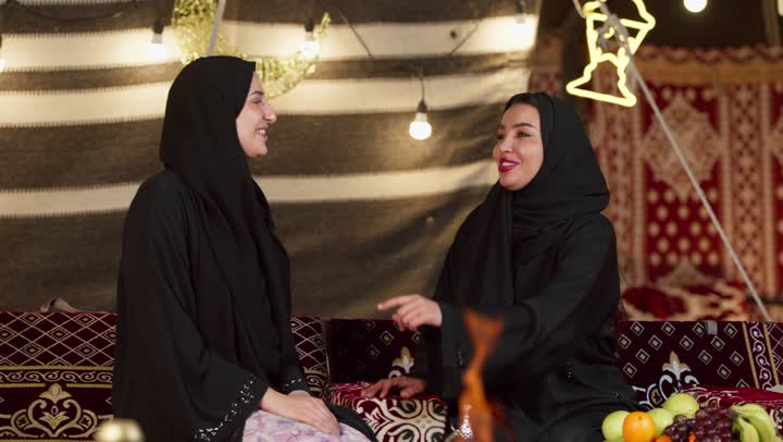 الإشارة بإصبع السبابة إلى شيء ما، حسن الاستقبال وكرم الضيافة في شهر رمضان, زينة وإضاءة رمضانية، كشتة بر وتخييم في صحراء الإمارات، حوار ونقاش بين الاصدقاء بإيماءات السعادة، سيدتان عربيتان اماراتيتان خليجيتان ترتديان العباءة تجلسان في خيمة شعبية