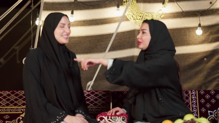 الإشارة بإصبع السبابة إلى شيء ما، حسن الاستقبال وكرم الضيافة في شهر رمضان, زينة وإضاءة رمضانية، كشتة بر وتخييم في صحراء الإمارات، حوار ونقاش بين الاصدقاء بإيماءات السعادة، سيدتان عربيتان اماراتيتان خليجيتان ترتديان العباءة تجلسان في خيمة شعبية