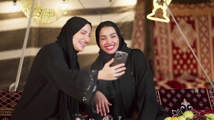استخدام جهاز تقني حديث، أجواء ليلية هادئة أمام موقد الحطب، زينة وإضاءة رمضانية، كشتة بر وتخييم في صحراء الإمارات، سيدتان عربيتان اماراتيتان خليجيتان ترتديان العباءة تجلسان في خيمة شعبية تقليدية تقومان بإجراء مكالمة فيديو عبر الهاتف المحمول بإيماءات السعادة