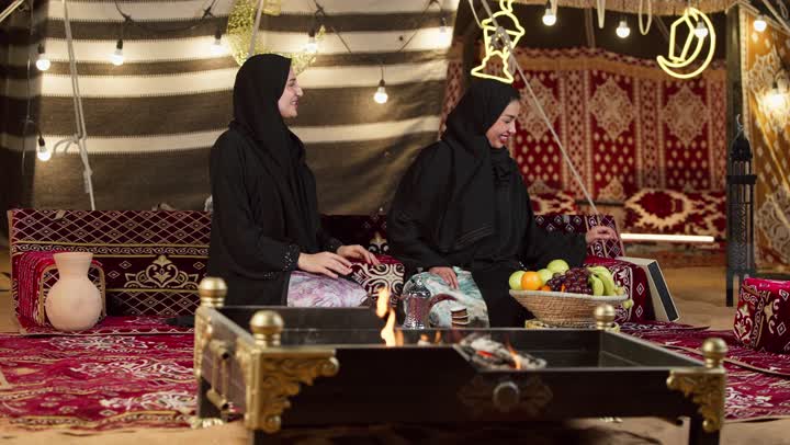 كشتة بر وتخييم في صحراء الإمارات، تقديم الهدية في المناسبات الدينية، قضاء وقت ممتع مع الأصدقاء، كرم الضيافة وحسن الاستقبال في شهر رمضان, سيدتان عربيتان اماراتيتان خليجيتان ترتديان العباءة تجلسان في خيمة شعبية تقليدية أمام موقد الحطب, زينة وإضاءة رمضانية