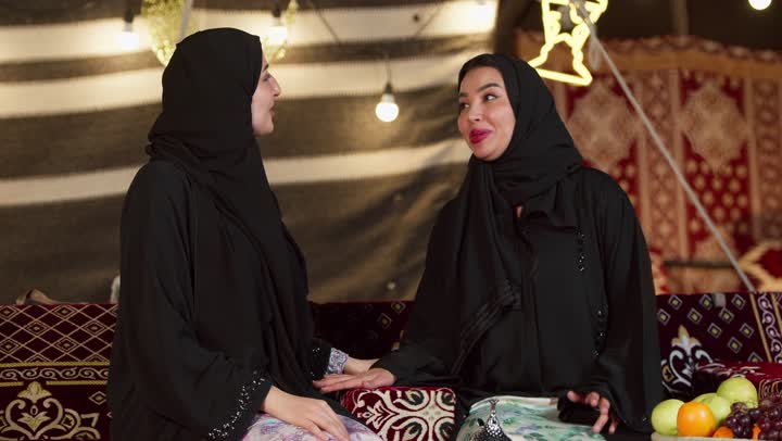 أجواء ليلية هادىْه في كشتة بر وتخييم في صحراء الإمارات، تقديم الهدية في المناسبات الدينية، قضاء وقت ممتع مع الأصدقاء، كرم الضيافة وحسن الاستقبال في شهر رمضان, سيدتان عربيتان اماراتيتان خليجيتان ترتديان العباءة تجلسان في خيمة شعبية تقليدية, ديكور وزينة وإضاءة رمضانية