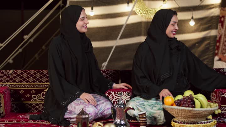 تقديم الهدية في المناسبات الدينية، قضاء وقت ممتع مع الأصدقاء، كرم الضيافة وحسن الاستقبال في شهر رمضان, سيدتان عربيتان اماراتيتان خليجيتان ترتديان العباءة تجلسان في خيمة شعبية تقليدية, ديكور وزينة وإضاءة رمضانية, أجواء ليلية هادىْه في كشتة بر وتخييم في صحراء الإمارات