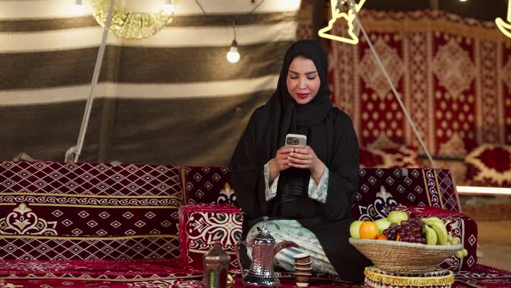 الكرم وحسن الضيافة عند العرب، كشتة بر وتخييم في صحراء الامارات، ديكور وزينة شهر رمضان المبارك، التواصل مع الأهل والاصدقاء عبر الجهاز التقني الحديث, سيدة عربية خليجية إماراتية محجبة ترتدي عباءة تقوم باستخدام الهاتف المحمول بإيماءات السعادة والسرور