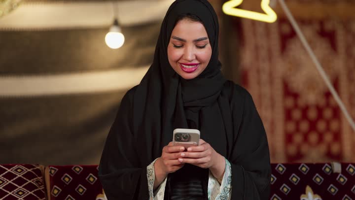 كشتة بر وتخييم في صحراء الامارات، ديكور وزينة شهر رمضان المبارك، التواصل مع الأهل والاصدقاء عبر الجهاز التقني الحديث, تصوير مقرب لسيدة عربية خليجية إماراتية محجبة ترتدي عباءة تقوم باستخدام الهاتف المحمول بإيماءات السعادة والسرور