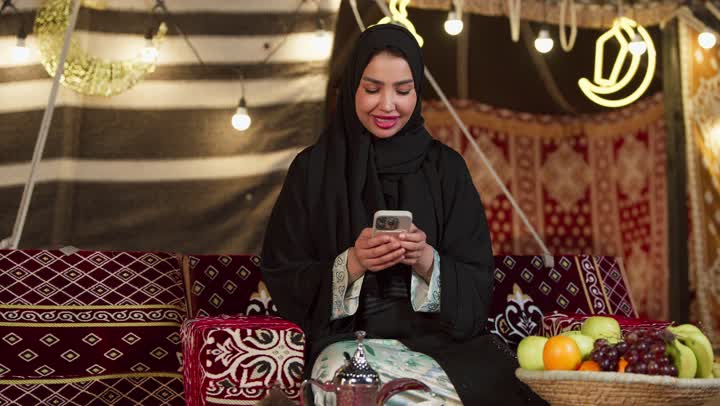 الكرم وحسن الضيافة عند العرب، كشتة بر وتخييم في صحراء الامارات، ديكور وزينة شهر رمضان المبارك، التواصل مع الأهل والاصدقاء عبر الجهاز التقني الحديث, سيدة عربية خليجية إماراتية محجبة ترتدي عباءة تقوم باستخدام الهاتف المحمول بإيماءات السعادة والسرور