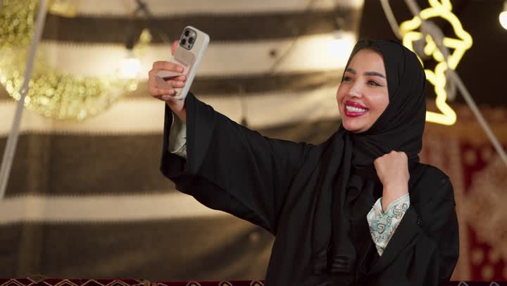 الاحتفاظ بالصور التذكارية من خلال الجهاز التقني الحديث, تصوير مقرب لسيدة عربية خليجية إماراتية محجبة ترتدي عباءة تقوم بالتقاط صورة سيلفي باستخدام الهاتف المحمول بتعابير السرور والسعادة، كشته بر وتخييم في صحراء الامارات, أجواء ليلية هادئة في خيمه شعبيه