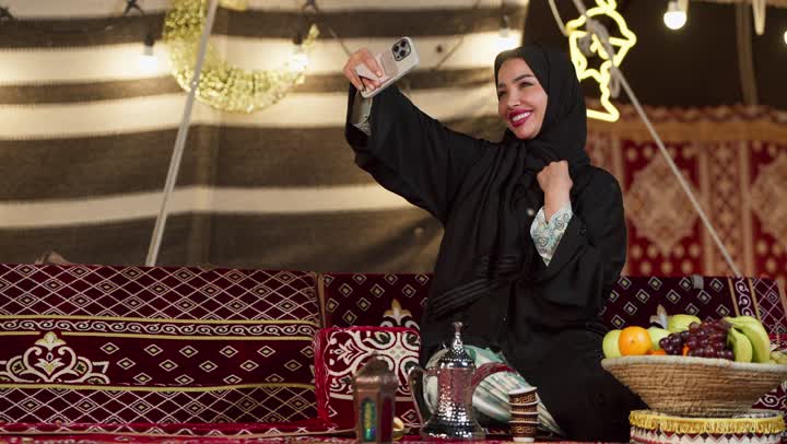 ديكور وزينة شهر رمضان المبارك، التواصل مع الأهل والاصدقاء عبر الجهاز التقني الحديث, سيدة عربية خليجية إماراتية محجبة ترتدي عباءة تقوم بإجراء مكالمة فيديو باستخدام الهاتف المحمول بإيماءات السعادة، كشته بر وتخييم في صحراء الامارات، إلقاء التحية والسلام