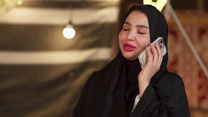 التواصل مع الأهل والاصدقاء عبر الجهاز التقني الحديث, كشتة بر وتخييم في صحراء الامارات، ديكور وزينة شهر رمضان المبارك، تصوير مقرب لسيدة عربية خليجية إماراتية محجبة ترتدي عباءة تقوم بإجراء مكالمة هاتفية عبر الهاتف المحمول بإيماءات السعادة