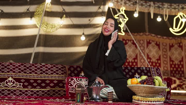 حسن وكرم الضيافة عند العرب، التواصل مع الأهل والاصدقاء عبر الجهاز التقني الحديث, كشتة بر وتخييم في صحراء الامارات، ديكور وزينة شهر رمضان المبارك، سيدة عربية خليجية إماراتية محجبة ترتدي عباءة تقوم بإجراء مكالمة هاتفية عبر الهاتف المحمول بإيماءات السعادة