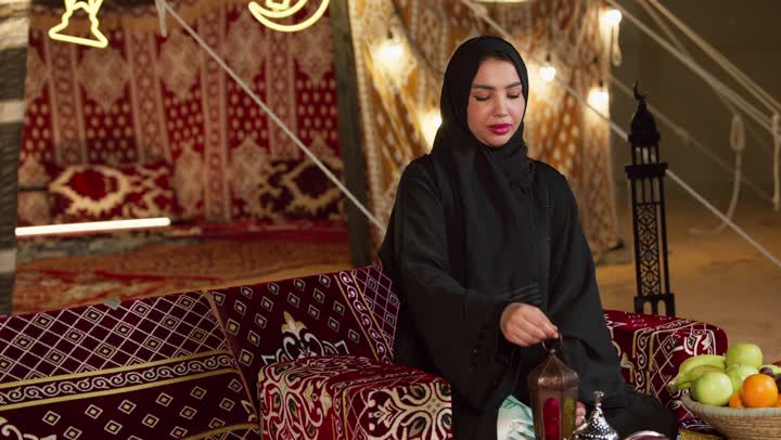 الإستعداد لقدوم شهر رمضان المبارك، أجواء السكينة والروحانية، الاحتفال بالمناسبات الدينية، سيدة عربية خليجية اماراتية محجبة ترتدي العباءة السوادء تجلس في خيمة شعبية وتحمل بيدها فانوس معدني، النظر الى الكاميرا بإيماءات السعادة والسرور، حسن الضيافة والكرم عند العرب