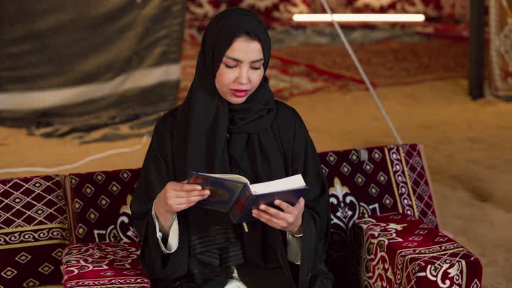 قراءة السورالقصيرة والورد اليومي، تلاوة آيات من القرآن الكريم، الاجتهاد في العبادة للتقرب إلى الله تعالى, كشتة بر وتخييم في صحراء الامارات، ديكور وزينة شهر رمضان المبارك، سيدة عربية خليجية إماراتية محجبة ترتدي عباءة تحمل بيدها المصحف الشريف وتقرأ منه