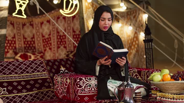 تلاوة آيات من القرآن الكريم، قراءة السورالقصيرة والورد اليومي، الاجتهاد في العبادة للتقرب إلى الله تعالى, كشتة بر وتخييم في صحراء الامارات، ديكور وزينة شهر رمضان المبارك، سيدة عربية خليجية إماراتية محجبة ترتدي عباءة تحمل بيدها المصحف الشريف وتقرأ منه