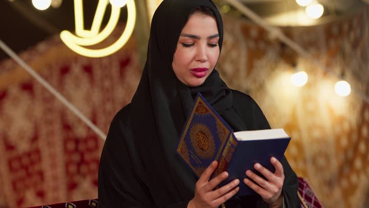 تلاوة آيات من القرآن الكريم، الاجتهاد في العبادة للتقرب إلى الله تعالى, الشعور بالخشوع والطمأنينة، قراءة السورالقصيرة والورد اليومي، كشتة بر وتخييم في صحراء الامارات، تصوير مقرب لسيدة عربية خليجية إماراتية ترتدي عباءة تحمل بيدها المصحف الشريف وتقرأ منه