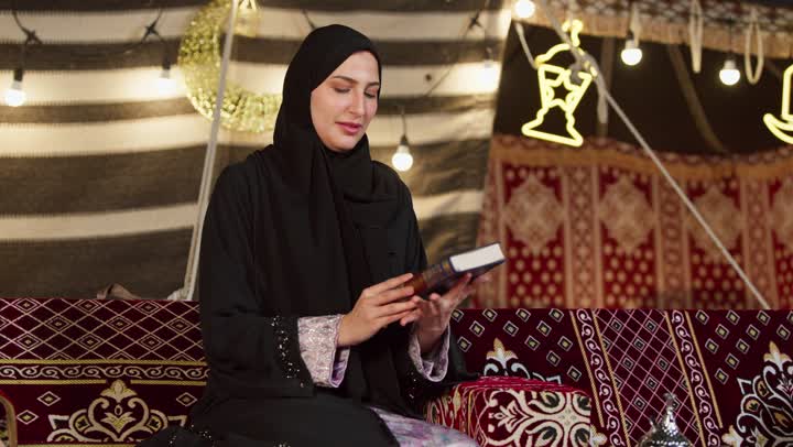قراءة السورالقصيرة والورد اليومي، تلاوة آيات من القرآن الكريم، الاجتهاد في العبادة للتقرب إلى الله تعالى, كشتة بر وتخييم في صحراء الامارات، ديكور وزينة شهر رمضان المبارك، سيدة عربية خليجية إماراتية محجبة ترتدي عباءة تحمل بيدها المصحف الشريف وتقرأ منه