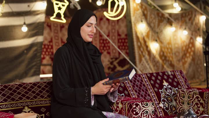 قراءة السورالقصيرة والورد اليومي، تلاوة آيات من القرآن الكريم، الاجتهاد في العبادة للتقرب إلى الله تعالى, كشتة بر وتخييم في صحراء الامارات، ديكور وزينة شهر رمضان المبارك، سيدة عربية خليجية إماراتية محجبة ترتدي عباءة تحمل بيدها المصحف الشريف وتقرأ منه