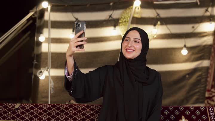 الاحتفاظ بالصور التذكارية من خلال الجهاز التقني الحديث, تصوير مقرب لسيدة عربية خليجية إماراتية محجبة ترتدي عباءة تقوم بالتقاط صورة سيلفي باستخدام الهاتف المحمول بتعابير السرور والسعادة، كشته بر وتخييم في صحراء الامارات, أجواء ليلية هادئة في خيمه شعبيه