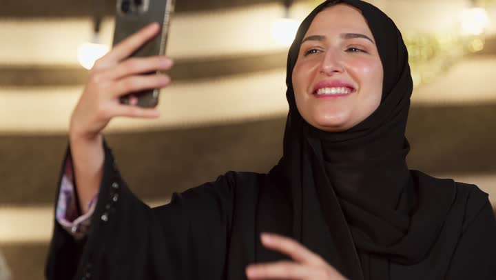 الاحتفاظ بالصور التذكارية من خلال الجهاز التقني الحديث, تصوير مقرب لسيدة عربية خليجية إماراتية محجبة ترتدي عباءة تقوم بالتقاط صورة سيلفي باستخدام الهاتف المحمول بتعابير السرور والسعادة، كشته بر وتخييم في صحراء الامارات, أجواء ليلية هادئة في خيمه شعبيه