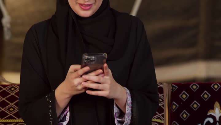 استخدام وسائل التواصل الاجتماعي عبر الجهاز التقني الحديث, كشتة بر وتخييم في صحراء الامارات، ديكور وزينة شهر رمضان المبارك، تصوير مقرب لسيدة عربية خليجية إماراتية محجبة ترتدي عباءة تقوم باستخدام الهاتف المحمول بإيماءات السعادة والسرور