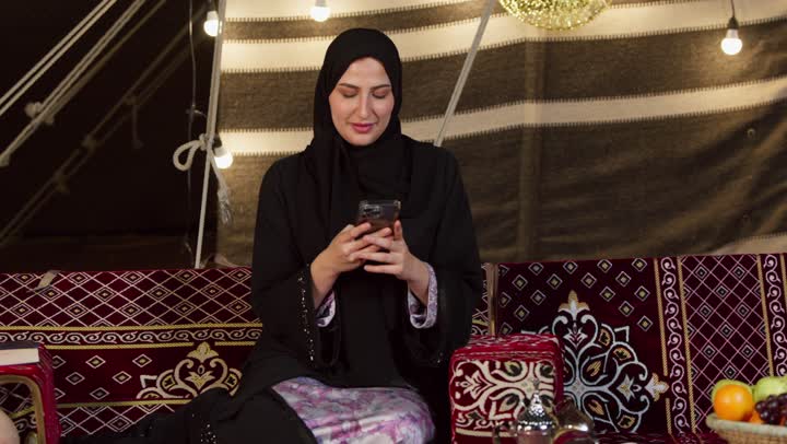 الكرم وحسن الضيافة عند العرب، كشتة بر وتخييم في صحراء الامارات، ديكور وزينة شهر رمضان المبارك، التواصل مع الأهل والاصدقاء عبر الجهاز التقني الحديث, سيدة عربية خليجية إماراتية محجبة ترتدي عباءة تقوم باستخدام الهاتف المحمول بإيماءات السعادة والسرور